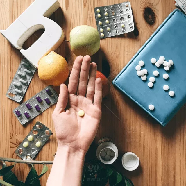 Pharmacology Homework Help: Comprehensive Guide Amidst Medications and Healthy Choices A hand holding a single tablet is surrounded by various pill blister packs, a prescription bottle, fruits, and a blue case on a wooden table—an ideal setting for anyone seeking pharmacology homework help